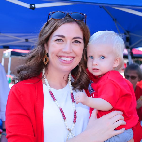 Woman wearing a read sweater with white shirt holding a young child wearing a red shirt in front of tailgate tents.